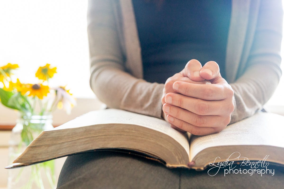 Hands clasped in prayer over Bible with bright light and flowers in background