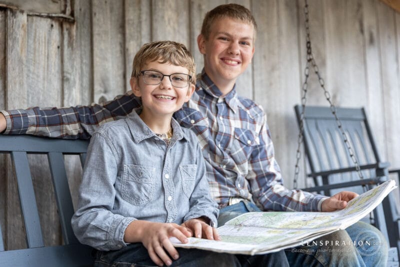 Two boys smile at the camera s they look at an atlas together on a swing on a porch