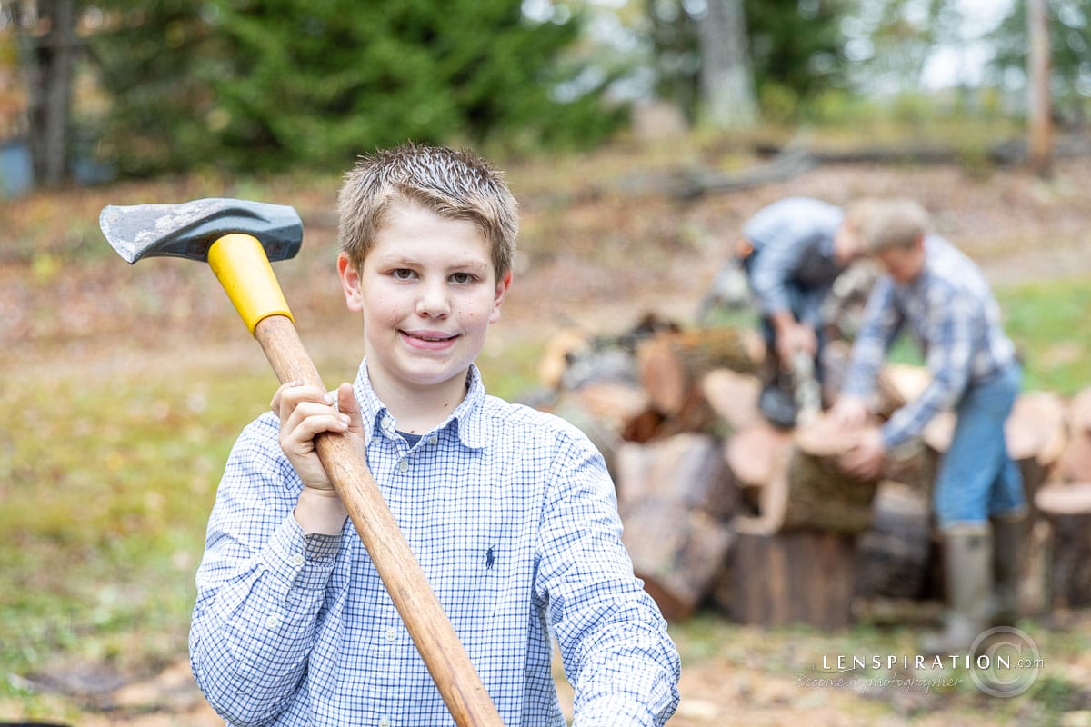 A young man holding a maul while his brothers work on a woodpile in the blurred background